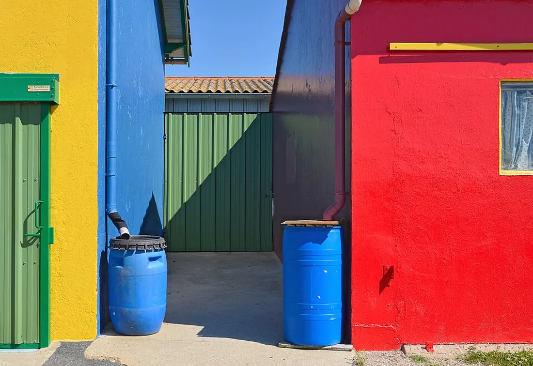Bidons bleus de réemploi raccordés aux gouttières de cabanes colorées pour récupérer l'eau de pluie